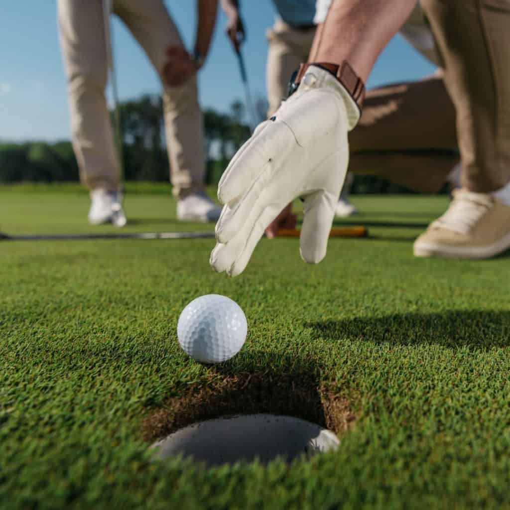 A golf ball on a golf course near a golf hole with a golfers hand about to push the golf ball into the golf hole with other golfers in the background.