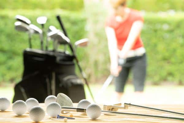 A bunch of golf balls and golf clubs on top of a wooden table with golf clubs in a golf bag and a female golfer in the background
