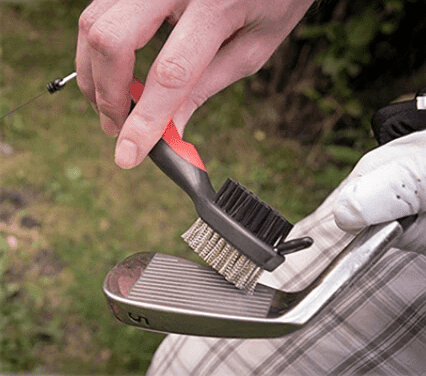 A golfer using a brush to clean a clubhead of a golf club while on the golf course