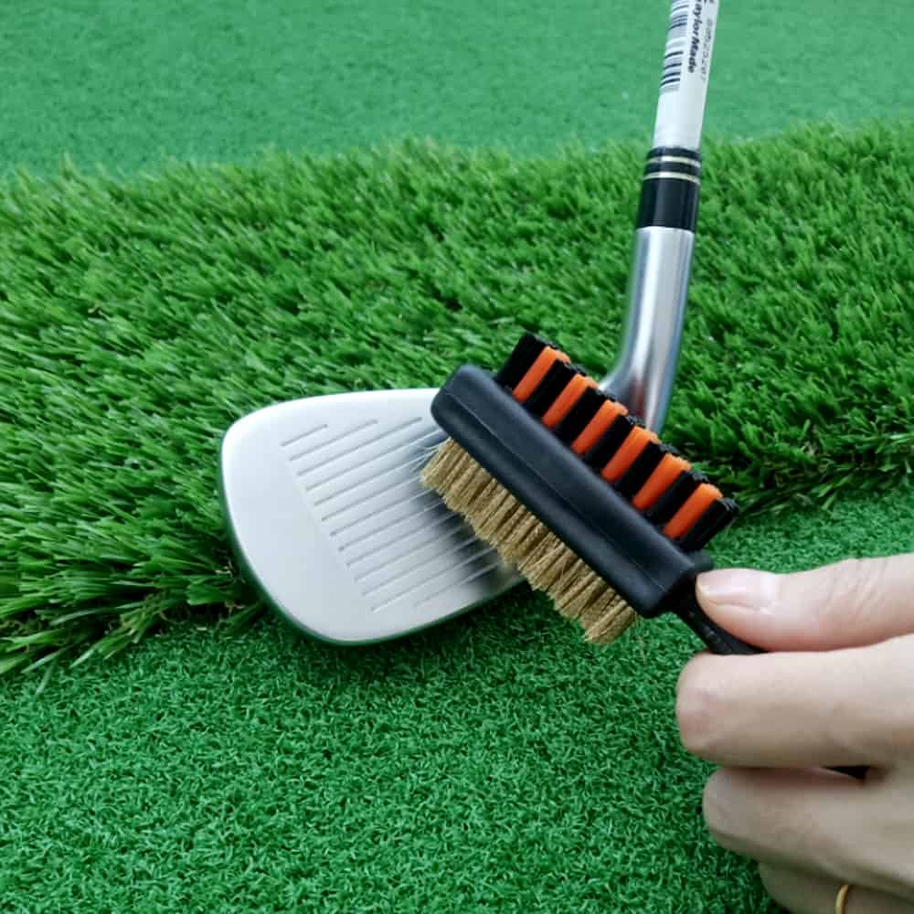 A person using a brush to clean the clubhead of a golf club on faux turf