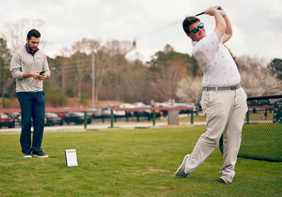 A golfer practicing his swing on the golf course with a launch monitor with his friend using his phone in the background