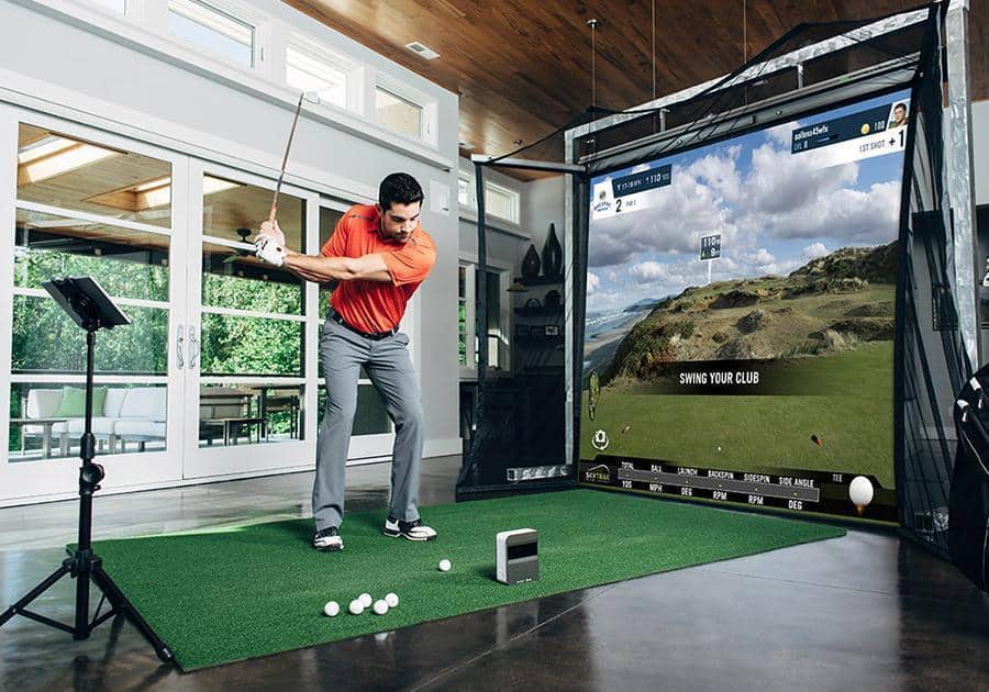Golfer playing on his indoor golf simulator setup complete with golf club, golf balls, hitting mat, projection screen, and other components