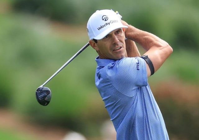 Billy Horschel in a blue shirt and white cap just after a swing using a hybrid golf club