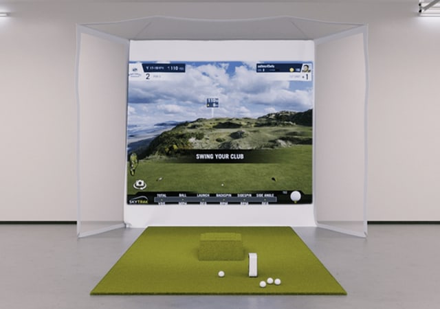 A SkyTrak Indoor Golf SImulator Flex Homecourse Package. Complete with net, projection screen, launch monitor, golf balls, and faux grass in a room.