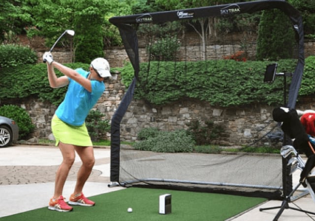 Woman swinging a golf club on a portable golf simulator setup in the outdoors. Golf net, launch monitors, and other golf accessories in the background.