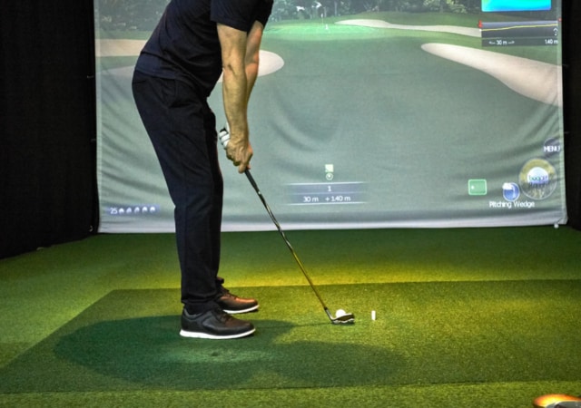 a golfer practicing his golf skills using a golf club and a golf ball in front of a golf simulator projection screen that shows a golf course simulation software