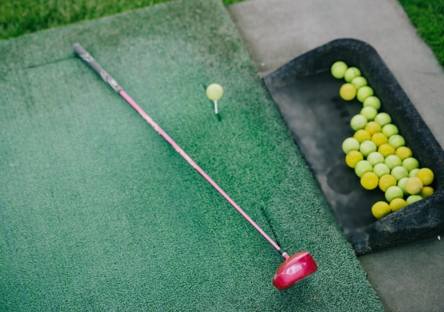 A red golf club placed on a golf hitting mat with a bunch of golf balls and a tee