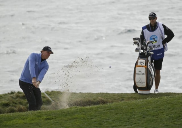 Phil Mickelson hitting out of a bunker with his golf bag and assistant in the background
