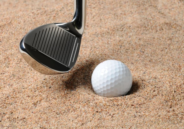 A golf ball in the sand with a golf club's head beside it