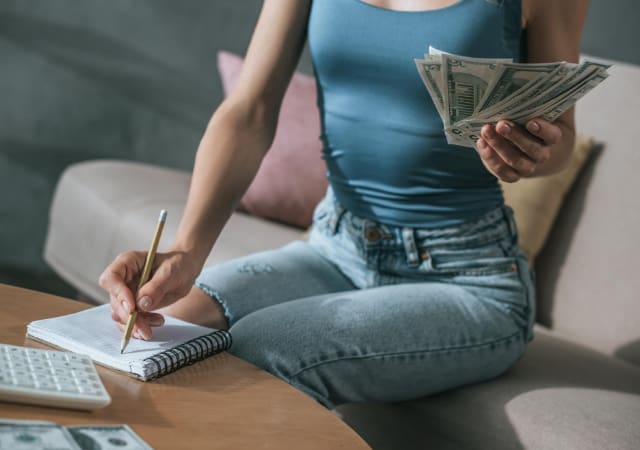 Woman sitting on a coach writing on a pad paper on the coffee table with one hand while holding money in the other
