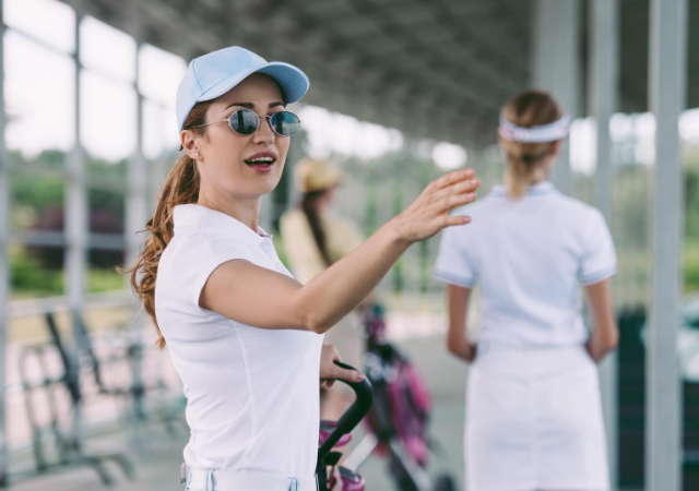 Female golfer talking to someone in a golf driving range wearing sunglasses and a hat