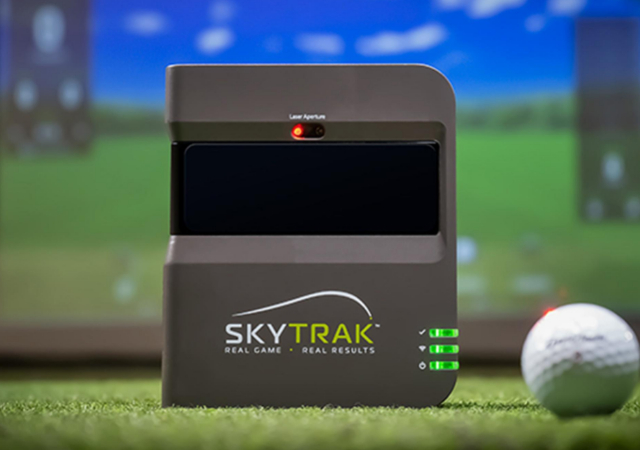 SkyTrak launch monitor with a golf ball beside it on turf with a projection screen at the back